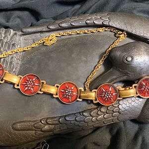 May include: A gold chain necklace with five red and gold round medallions. Each medallion features a stylized starburst design.
