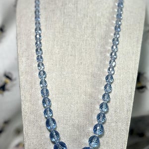 May include: A light blue beaded necklace displayed on a mannequin. The necklace features faceted, round beads of varying sizes, with larger beads clustered at the center. The beads have a translucent quality, reflecting light. The necklace is a vintage style.