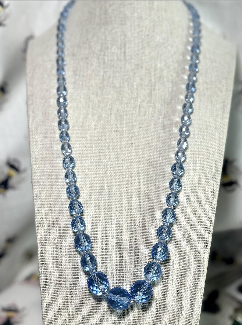 May include: A light blue beaded necklace displayed on a mannequin. The necklace features faceted, round beads of varying sizes, with larger beads clustered at the center. The beads have a translucent quality, reflecting light. The necklace is a vintage style.