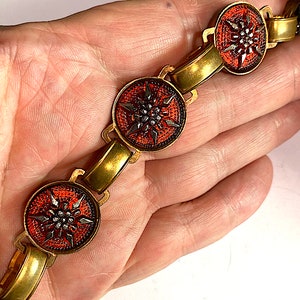 May include: A gold bracelet with red and black enamel flower charms. The flowers have a stylized design with a center of small black beads.