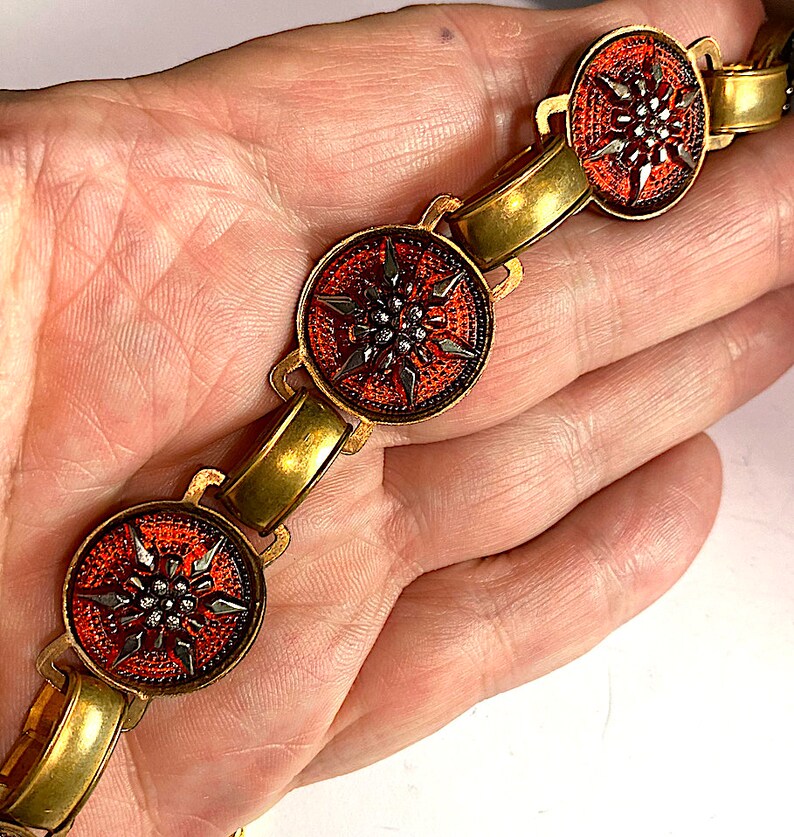 May include: A gold bracelet with red and black enamel flower charms. The flowers have a stylized design with a center of small black beads.