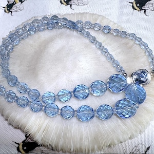 May include: A light blue beaded necklace with faceted beads of varying sizes. The necklace is displayed on a white, textured surface with a bee-themed fabric background. The necklace has a silver clasp.