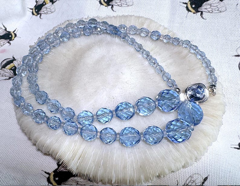 May include: A light blue beaded necklace with faceted beads of varying sizes. The necklace is displayed on a white, textured surface with a bee-themed fabric background. The necklace has a silver clasp.