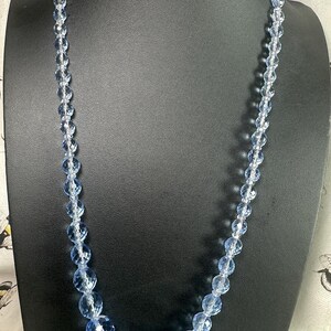 May include: A light blue beaded necklace with faceted, round beads of varying sizes. The necklace is displayed on a black mannequin. The beads have a clear, sparkling appearance, suggesting a glass or crystal material.