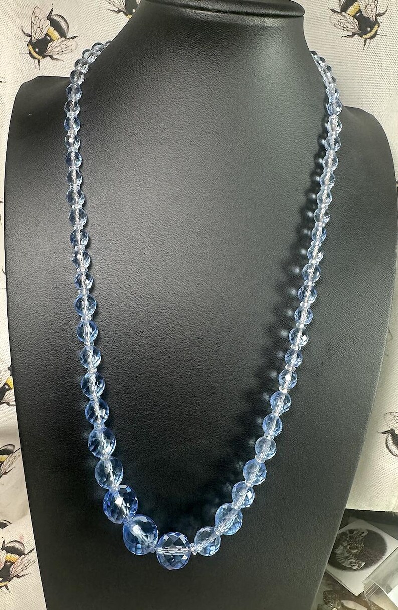 May include: A light blue beaded necklace with faceted, round beads of varying sizes. The necklace is displayed on a black mannequin. The beads have a clear, sparkling appearance, suggesting a glass or crystal material.