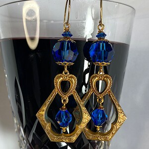 K&ouml;nnte beinhalten: Goldfarbene H&auml;ngeohrringe mit facettierten blauen Kristallperlen. Die Ohrringe haben ein herzf&ouml;rmiges Design und einen rautenf&ouml;rmigen Rahmen mit floralen Details. Die Ohrringe sind an goldfarbenen Ohrhaken befestigt.