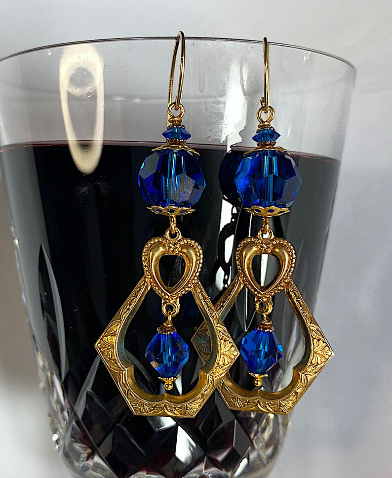 K&ouml;nnte beinhalten: Goldfarbene H&auml;ngeohrringe mit facettierten blauen Kristallperlen. Die Ohrringe haben ein herzf&ouml;rmiges Design und einen rautenf&ouml;rmigen Rahmen mit floralen Details. Die Ohrringe sind an goldfarbenen Ohrhaken befestigt.