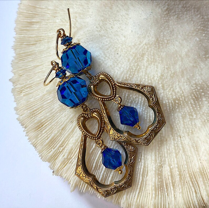 K&ouml;nnte beinhalten: Ein Paar verzierte goldfarbene Ohrringe mit blauen Kristallakzenten. Die Ohrringe haben ein geometrisches Design mit herzf&ouml;rmigen Ausschnitten und h&auml;ngenden blauen Kristallen. Sie werden auf einer wei&szlig;en, strukturierten Oberfl&auml;che pr&auml;sentiert.