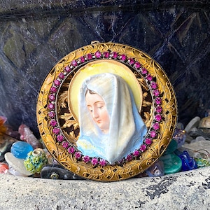 May include: A gold-toned pendant with a round, ornate frame featuring a cameo of a woman with a white veil and a blue robe. The frame is decorated with pink rhinestones.
