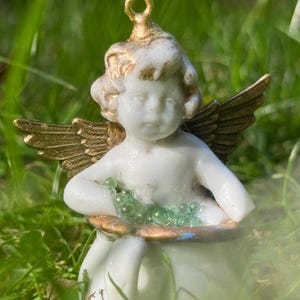 May include: A porcelain cherub figurine with gold wings and a green glass bead garland. The cherub is sitting on a white porcelain cup.