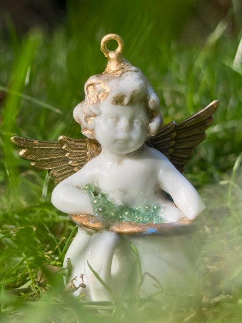 May include: A porcelain cherub figurine with gold wings and a green glass bead garland. The cherub is sitting on a white porcelain cup.