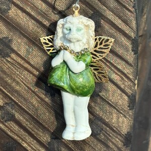 Puede incluir: Pequeña figura blanca de ángel con ojos verdes y vestido verde. El ángel tiene alas de filigrana doradas y un halo dorado. El ángel junta las manos en posición de oración. La figura está colgada de un gancho plateado.
