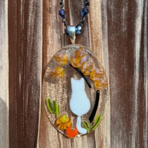 May include: A white glass pendant necklace with a white cat silhouette, orange and yellow flowers, and green leaves. The necklace is on a blue beaded chain.