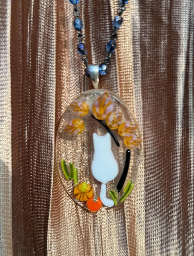 May include: A white glass pendant necklace with a white cat silhouette, orange and yellow flowers, and green leaves. The necklace is on a blue beaded chain.