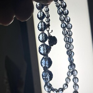 May include: A vintage necklace featuring faceted, dark blue beads. The necklace is composed of two strands of varying bead sizes, with a clasp closure. The beads have a reflective quality, catching the light.