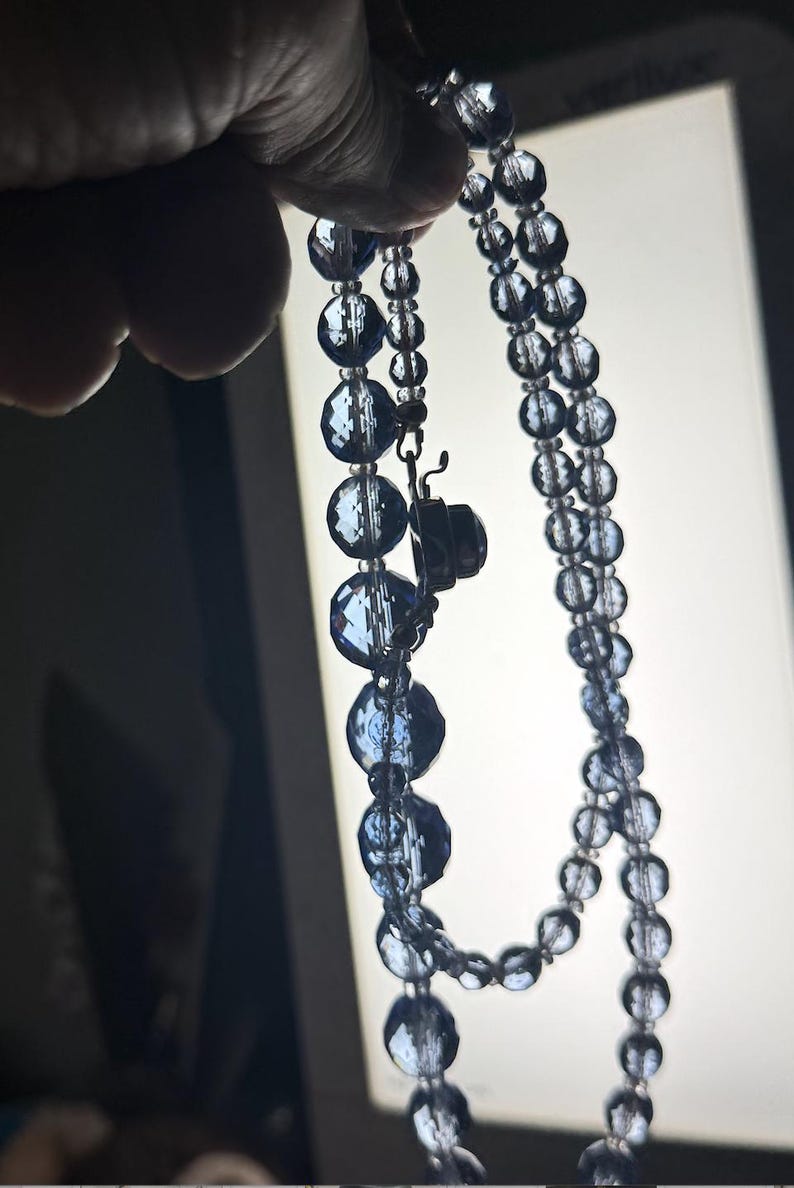 May include: A vintage necklace featuring faceted, dark blue beads. The necklace is composed of two strands of varying bead sizes, with a clasp closure. The beads have a reflective quality, catching the light.