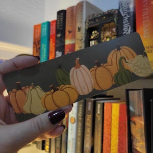 May include: A rectangular bookmark featuring a row of illustrated pumpkins in shades of orange, green, and white against a dark green background. The bookmark is held in front of a bookshelf filled with books.