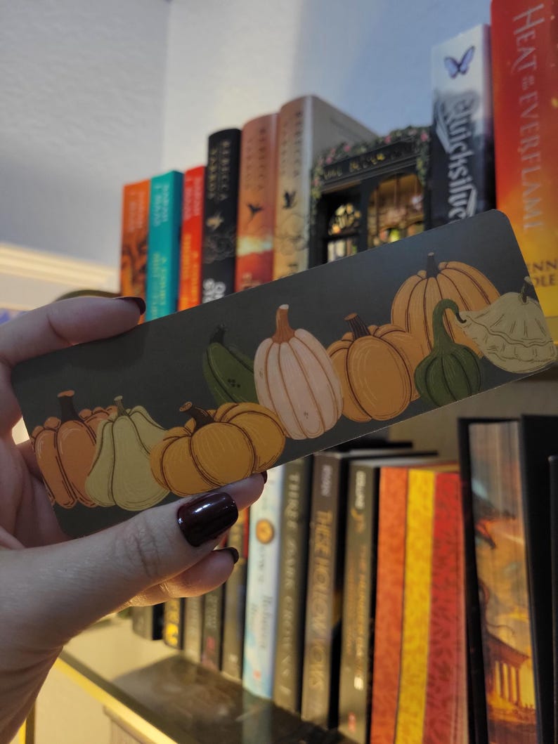 May include: A rectangular bookmark featuring a row of illustrated pumpkins in shades of orange, green, and white against a dark green background. The bookmark is held in front of a bookshelf filled with books.
