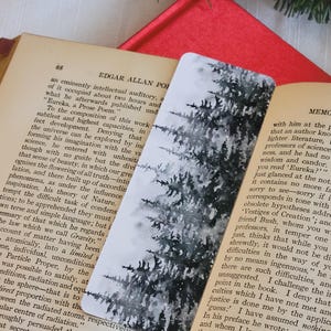 Könnte beinhalten: Rechteckiger Lesezeichen mit einem Aquarellgemälde eines verschneiten Nadelwaldes. Das Lesezeichen liegt auf den Seiten eines offenen Buches, mit einem roten Band im Hintergrund.