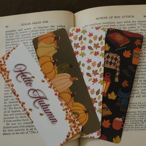 May include: Four decorative bookmarks with autumn-themed designs are displayed on an open book. The bookmarks feature pumpkins, fall leaves, and the phrase "Hello Autumn." The color palette includes orange, brown, and white.