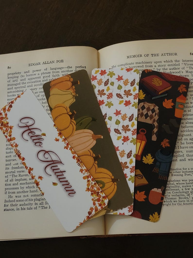 May include: Four decorative bookmarks with autumn-themed designs are displayed on an open book. The bookmarks feature pumpkins, fall leaves, and the phrase "Hello Autumn." The color palette includes orange, brown, and white.