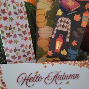 May include: A collection of autumn-themed bookmarks. One bookmark features a white background with colorful leaves. Another shows pumpkins, and a third displays a sweater, books, and a coffee cup. The bottom bookmark reads "Hello Autumn".