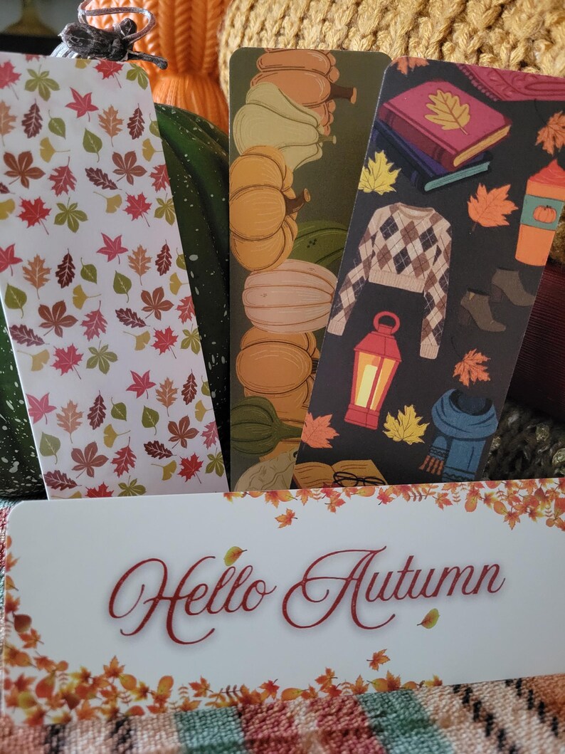 May include: A collection of autumn-themed bookmarks. One bookmark features a white background with colorful leaves. Another shows pumpkins, and a third displays a sweater, books, and a coffee cup. The bottom bookmark reads "Hello Autumn".