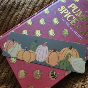 May include: A bookmark featuring a row of illustrated pumpkins in various colors, including orange, white, and green, against a teal background. The bookmark rests on a book with the title "Pumpkin Spice Cake" by Laurie Gilmore.