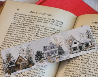 May include: A decorative bookmark featuring a winter scene with houses, trees, and snow. The houses are painted in shades of brown, gray, and white. The bookmark is resting on an open book.