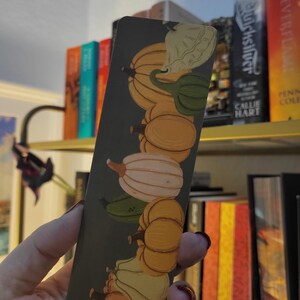 May include: A rectangular bookmark featuring a vertical arrangement of illustrated gourds in shades of orange, green, and white against a dark green background. The bookmark is held in a hand, with a bookshelf visible in the background.