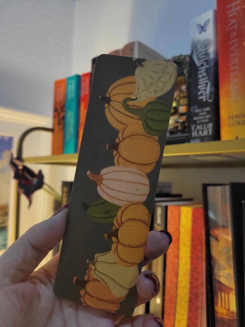 May include: A rectangular bookmark featuring a vertical arrangement of illustrated gourds in shades of orange, green, and white against a dark green background. The bookmark is held in a hand, with a bookshelf visible in the background.