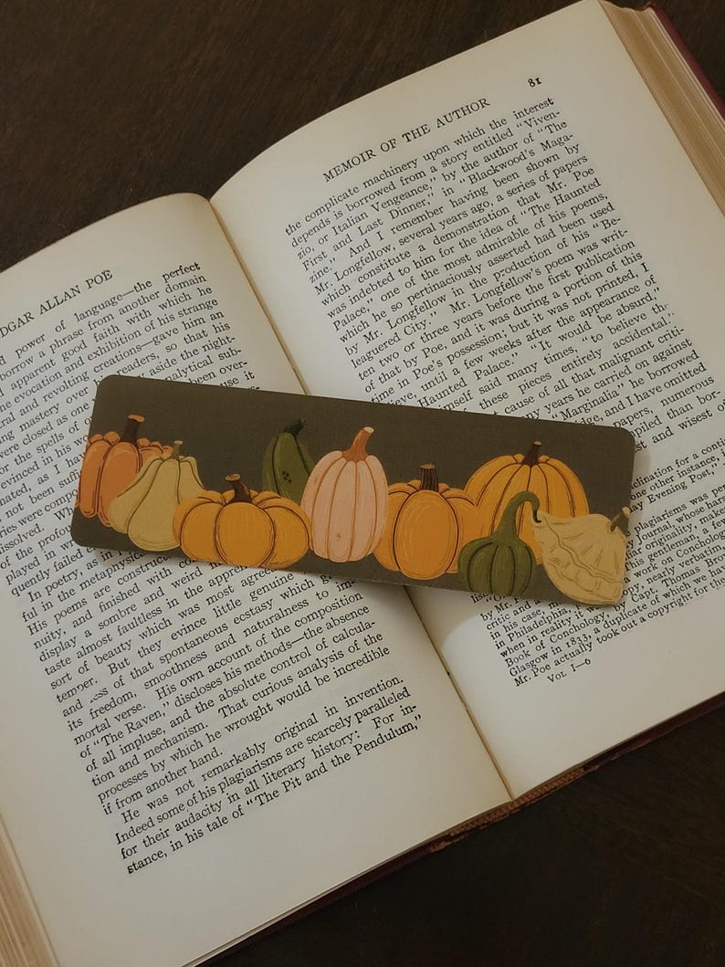 May include: A decorative bookmark featuring a fall harvest theme. The bookmark displays a series of pumpkins in various shapes, sizes, and colors, including orange, yellow, and green. The bookmark is placed on an open book.