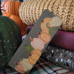 May include: A rectangular bookmark featuring illustrations of various pumpkins in shades of orange, white, and green against a gray background. The bookmark is positioned in front of a stack of books and a knitted pumpkin.