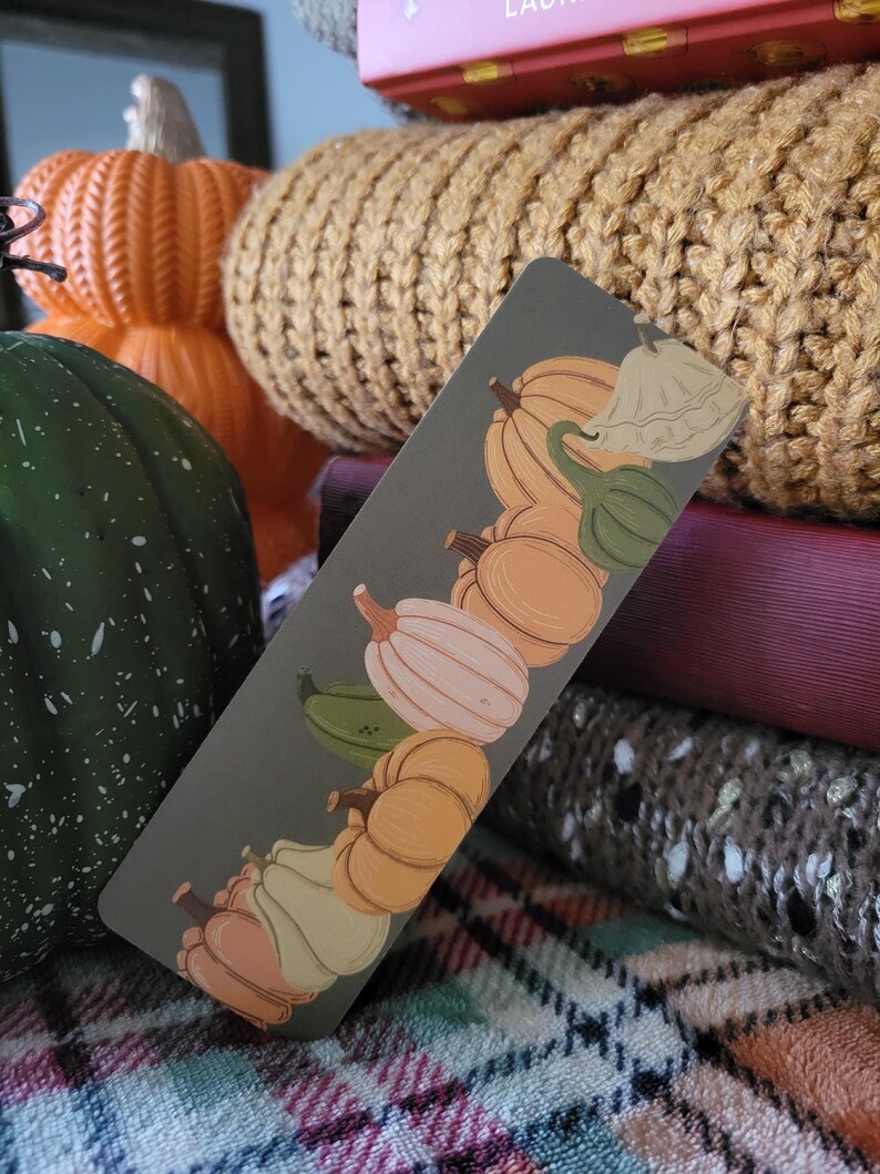 May include: A rectangular bookmark featuring illustrations of various pumpkins in shades of orange, white, and green against a gray background. The bookmark is positioned in front of a stack of books and a knitted pumpkin.