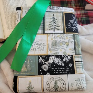 May include: A fabric book sleeve with a green ribbon bookmark. The sleeve features a patchwork design with Christmas-themed patterns, including Christmas trees and the text "Merry Christmas". The background includes an open book.