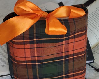 May include: A plaid fabric gift bag in orange, green, and brown, tied with a bright orange satin bow. The bag is rectangular and sits on a soft, white surface, with the edges of open books visible in the background.