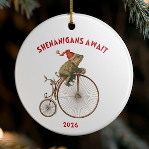 May include: A round white ceramic ornament featuring a green frog wearing a red Santa hat while riding a vintage penny-farthing bicycle. The text SHENANIGANS AWAIT is printed in red above the frog, and the year 2026 is printed in red below.