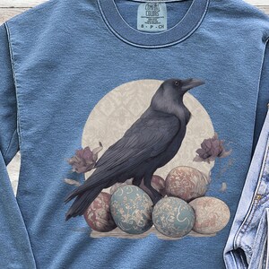 Puede incluir: Una sudadera azul de manga larga con cuello redondo que presenta un gráfico de un cuervo negro posado sobre un grupo de huevos estampados frente a un fondo circular beige. La sudadera tiene un cuello acanalado y una etiqueta de Comfort Colors.