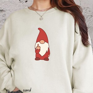 May include: A sand-colored crewneck sweatshirt featuring a graphic of a gnome wearing a red hat and beard, depicted with a middle finger gesture. The garment has long sleeves and a ribbed neckline, with the word sand printed near the bottom hem.