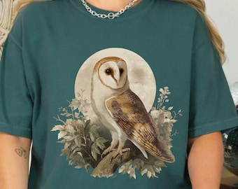 Puede incluir: Camiseta de color verde azulado con una ilustración detallada de un búho común posado en una rama frente a una luna llena. El búho tiene plumas marrones y blancas. Las palabras "blue spruce" están impresas en la parte inferior.