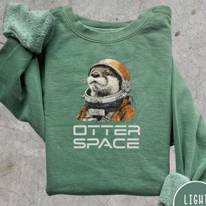 May include: Light green sweatshirt featuring an otter wearing an astronaut helmet and suit. The text "OTTER SPACE" is printed in white below the otter illustration. The sweatshirt has a crew neck and long sleeves.