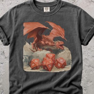 以下が含まれることがあります： 赤色のドラゴンと赤いサイコロのイラストが描かれた濃いグレーのTシャツ。ドラゴンは大きな翼を持ち、岩の上にいます。サイコロには数字が書かれています。シャツの下部には「pepper」の文字があります。