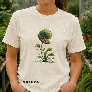Puede incluir: Camiseta beige con una ilustración detallada de una planta carnívora Venus. La planta tiene hojas verdes y un interior rojo. La palabra "NATURAL" está impresa en negro en la parte inferior de la camiseta.