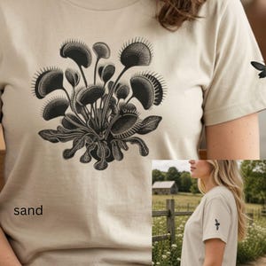 以下が含まれることがあります： サンドカラーのTシャツで、前面にハエトリグサの大きな黒いグラフィック、袖に小さな黒い昆虫のグラフィックが描かれています。シャツの左下には、sandという文字が小さな黒いフォントでプリントされています。
