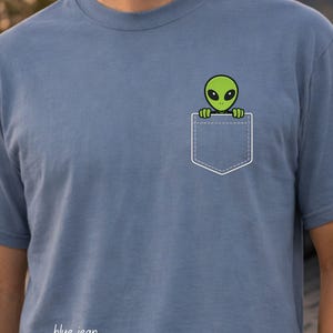 May include: Blue t-shirt with a white pocket design featuring a green alien peeking out. The text "blue jean" is printed at the bottom left of the shirt. The shirt is made of a soft material.