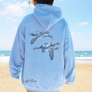 Op de afbeelding: Lichtblauwe hoodie met twee gedetailleerde illustraties van zeeschildpadden op de rug. De hoodie heeft een capuchon met trekkoord en lange mouwen. De tekst "Light Blue" staat onderaan gedrukt. De achtergrond toont een strand en een blauwe lucht.