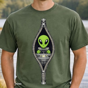 Puede incluir: Camiseta verde oliva con un gráfico de un alienígena verde conduciendo un vehículo dentro de una cremallera. El alienígena tiene grandes ojos negros y sostiene un volante. La cremallera es negra y plateada.