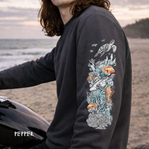 May include: A charcoal gray long-sleeve sweatshirt featuring a vertical graphic print on the sleeve. The design depicts sea turtles, orange fish, and coral reef elements in a detailed illustration style. The word PEPPER is printed in white.