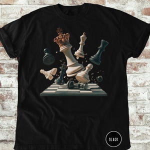 May include: Black t-shirt featuring a dynamic chess scene with pieces seemingly exploding from the board. The design includes a white king with a gold crown, white and black chess pieces, and a checkered board. The word "BLACK" is printed on the bottom right.