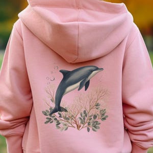 Puede incluir: Sudadera con capucha rosa claro con un diseño de delfín. La ilustración muestra un delfín gris y blanco saltando sobre corales y burbujas. La sudadera con capucha está hecha de un material suave.